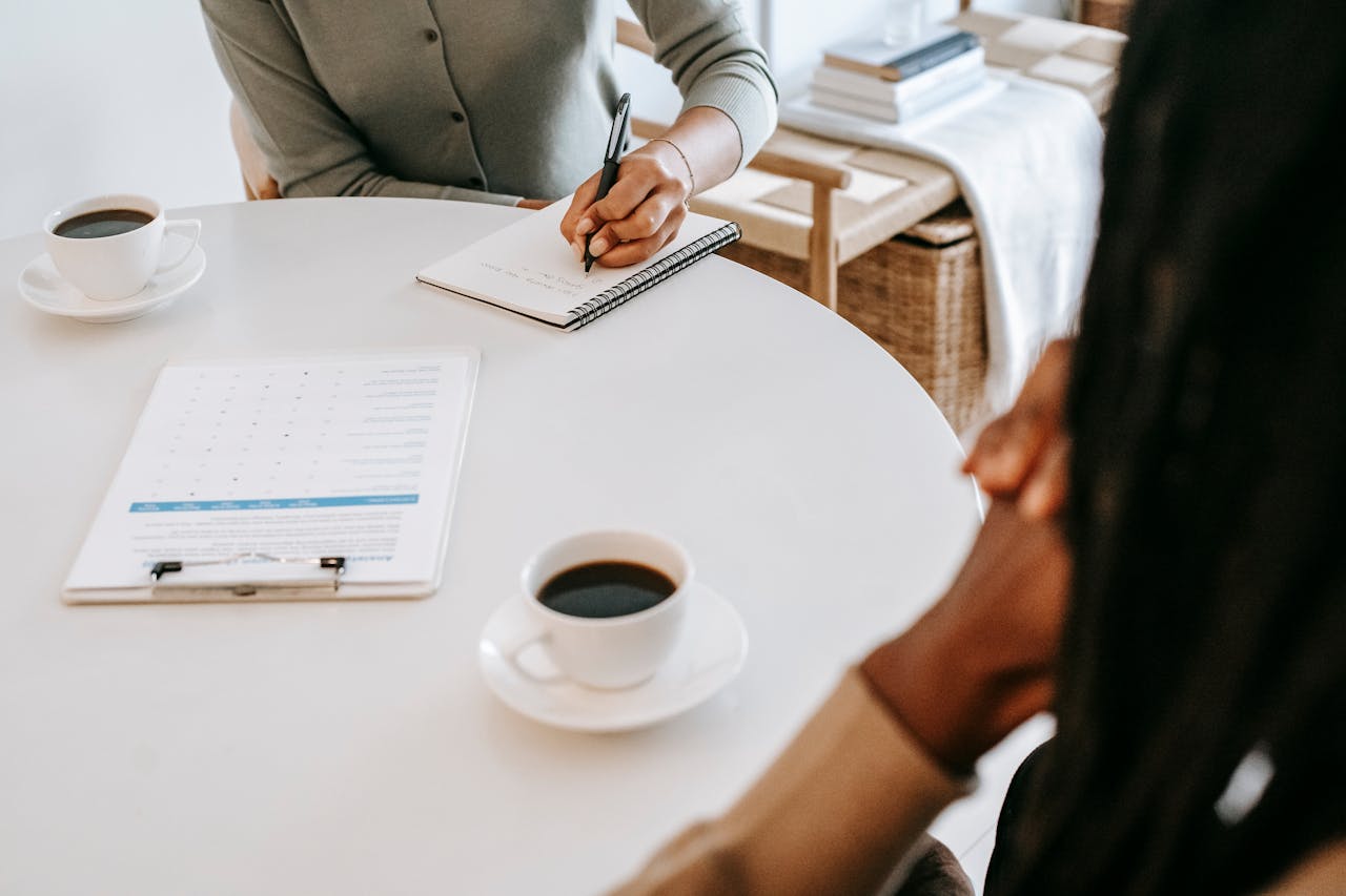 our-services-1 From above crop multiracial female interviewer or psychologist in formal wear asking questions and taking notes in planner while talking to black man and sitting together at table with coffee